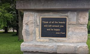 An image of a quote by Anne Frank at a rock in Columbus Park of Roses. The quote says: "Think of all the beauty still left around you and be happy."