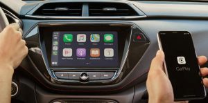 The dashboard control center of a 2023 Chevrolet Trailblazer with one hand driving from the driver's seat, and another holding her iphone displaying Apple Carplay from the passenger seat.