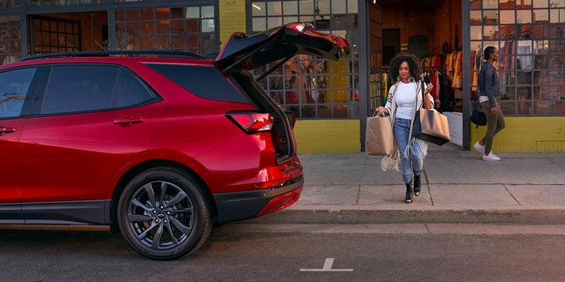 A red 2024 Chevrolet Equinox parked on a curb with the trunk open. The owner is walking towards the back of the car with bags to pack away.