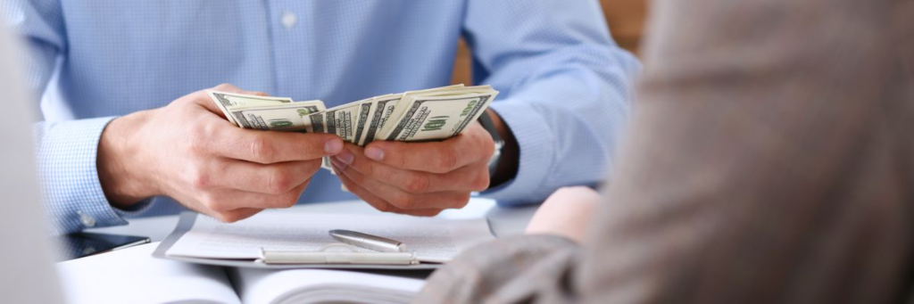 A person counting stacks of hundred-dollar bills on a desk with a pen and paperwork.